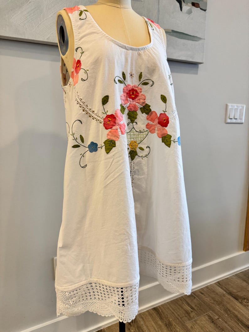 May include: White sleeveless dress with floral embroidery in pink, red, and green. The dress features a scoop neckline and a crochet lace hem. The floral design is centered on the front, with additional embroidery along the neckline.