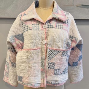 May include: A quilted jacket in a patchwork design, featuring a mix of white, pink, and blue fabrics. The jacket has a collared neckline, two front pockets, and large white buttons. The edges are trimmed with pink piping.