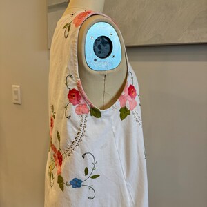 May include: White sleeveless dress with floral embroidery in pink, red, and blue. The dress features a scoop neckline and a relaxed fit. The embroidery includes flowers and vines, adding a touch of color and detail to the garment. The dress is displayed on a mannequin.