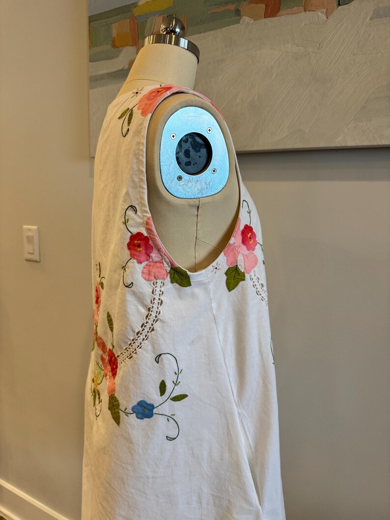 May include: White sleeveless dress with floral embroidery in pink, red, and blue. The dress features a scoop neckline and a relaxed fit. The embroidery includes flowers and vines, adding a touch of color and detail to the garment. The dress is displayed on a mannequin.