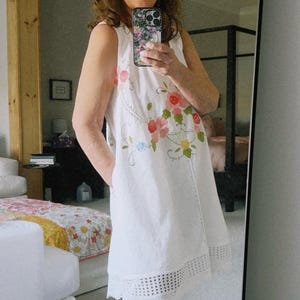 May include: White sleeveless dress with floral embroidery and a scalloped hem. The dress is paired with light blue cowboy boots.