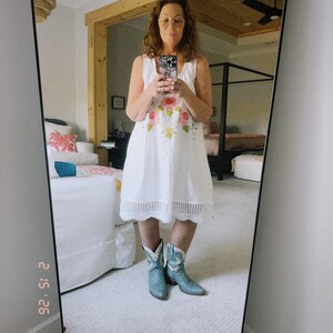 May include: A white sleeveless dress with floral embroidery and a scalloped hem is paired with light blue cowboy boots. The image is a mirror selfie, reflecting a bedroom setting with a bed and a ceiling fan.