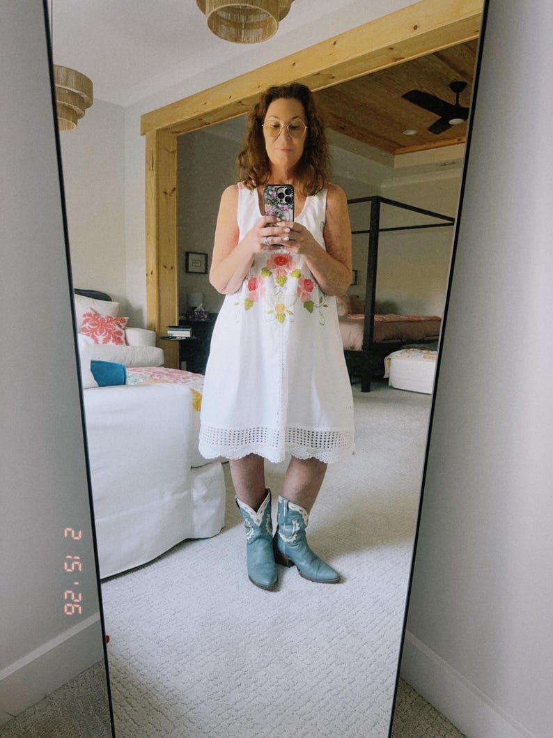 May include: A white sleeveless dress with floral embroidery and a scalloped hem is paired with light blue cowboy boots. The image is a mirror selfie, reflecting a bedroom setting with a bed and a ceiling fan.