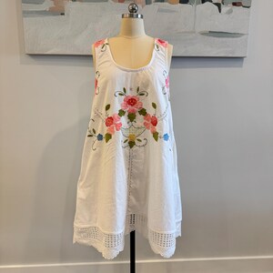 May include: White sleeveless dress with floral embroidery in pink, red, and blue. The dress features a scoop neckline and a lace trim at the hem. The dress is displayed on a mannequin.