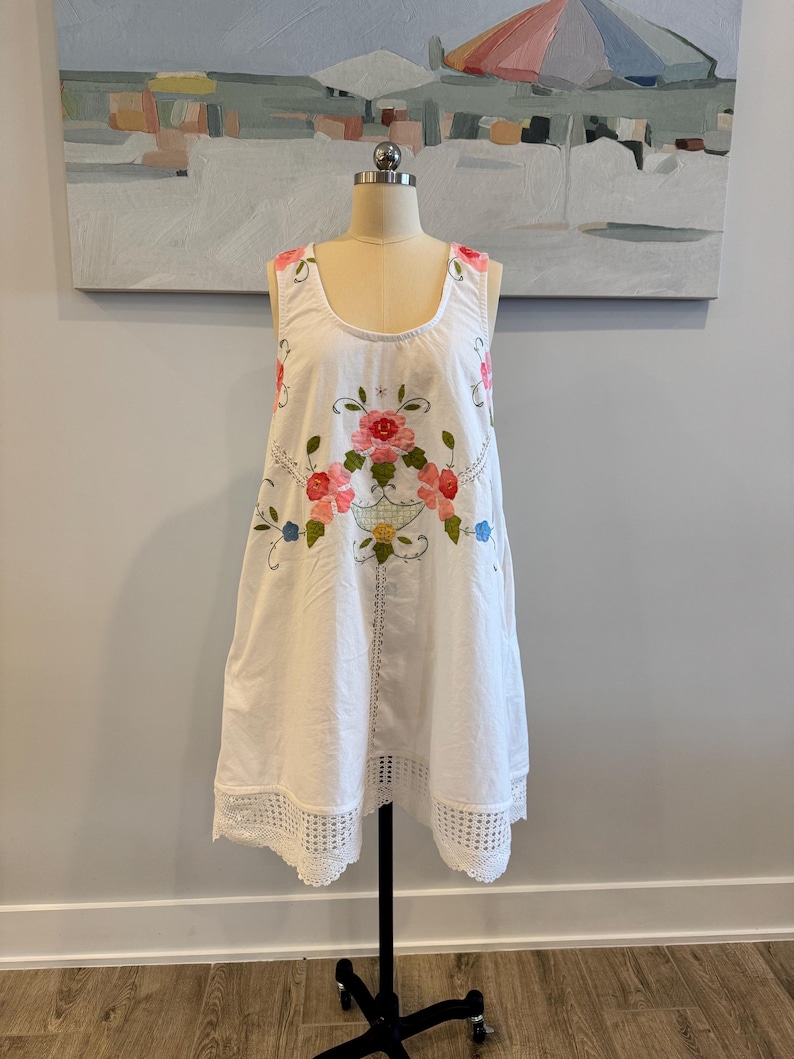 May include: White sleeveless dress with floral embroidery in pink, red, and blue. The dress features a scoop neckline and a lace trim at the hem. The dress is displayed on a mannequin.