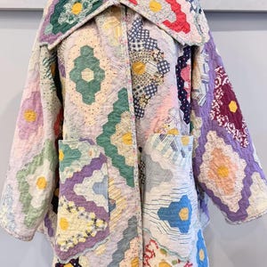 May include: A long-sleeved jacket constructed from a patchwork quilt featuring a hexagonal pattern. The garment displays a variety of floral and geometric fabric prints in shades of green, purple, red, blue, and yellow, with two large front pockets.