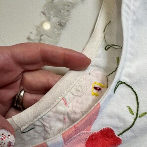 May include: Close-up of a white fabric with floral embroidery. The embroidery features red, pink, and yellow flowers with green stems and leaves. The edge of the fabric is trimmed with a cream-colored binding.