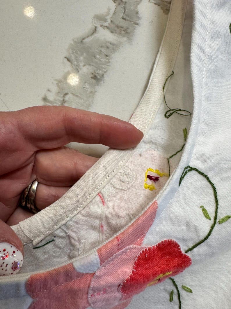 May include: Close-up of a white fabric with floral embroidery. The embroidery features red, pink, and yellow flowers with green stems and leaves. The edge of the fabric is trimmed with a cream-colored binding.