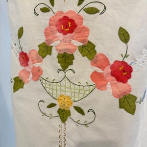 May include: White fabric with a floral appliqu&eacute; design. The design features pink and red flowers with green leaves, arranged in a basket-like pattern. Delicate lace trim and embroidered details add to the handcrafted aesthetic.