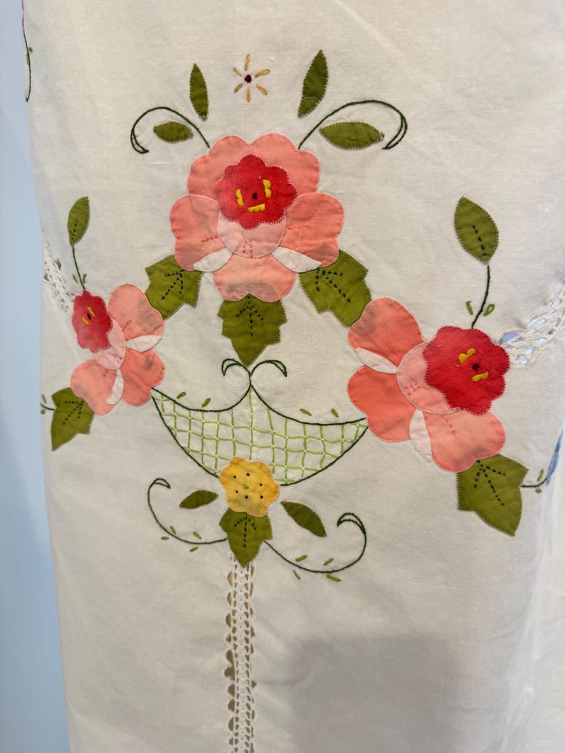May include: White fabric with a floral appliqu&eacute; design. The design features pink and red flowers with green leaves, arranged in a basket-like pattern. Delicate lace trim and embroidered details add to the handcrafted aesthetic.