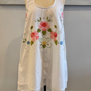 May include: White sleeveless dress with a floral embroidery design in pink, red, green, and blue. The dress features a scoop neckline, a crochet lace hem, and a decorative floral basket design.