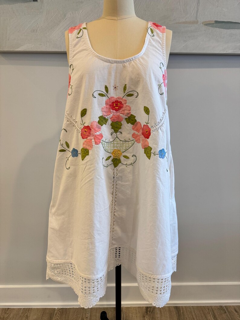 May include: White sleeveless dress with a floral embroidery design in pink, red, green, and blue. The dress features a scoop neckline, a crochet lace hem, and a decorative floral basket design.