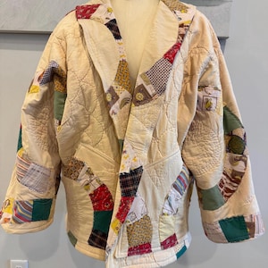 May include: Cream-colored quilted jacket with a patchwork design. The jacket features a notched collar and is adorned with various fabric patches in red, green, yellow, and patterned fabrics. The jacket is displayed on a mannequin.