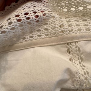 May include: Close-up of off-white fabric with a crocheted lace trim. The lace features an open, geometric pattern. The fabric appears to be a soft, woven material. The image highlights the texture and detail of the textile.