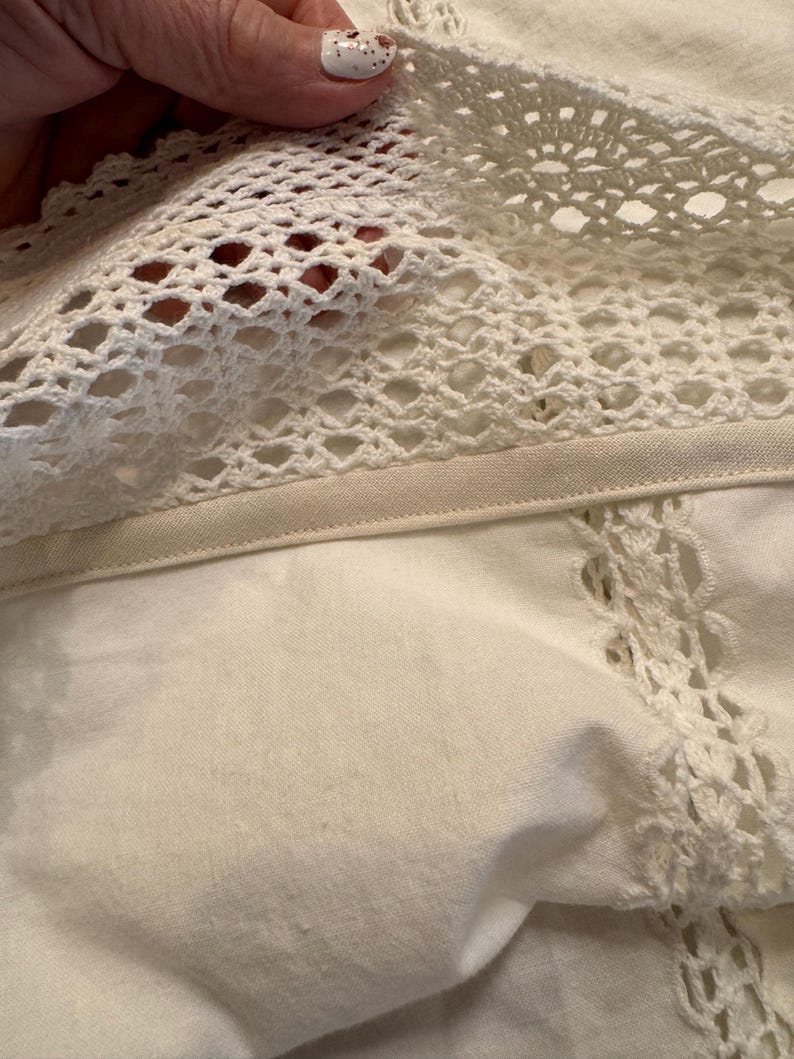 May include: Close-up of off-white fabric with a crocheted lace trim. The lace features an open, geometric pattern. The fabric appears to be a soft, woven material. The image highlights the texture and detail of the textile.