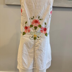 May include: White sleeveless dress with floral embroidery in pink, red, and green. The dress features a crochet lace hem and a decorative floral design on the back. The dress is displayed on a mannequin.