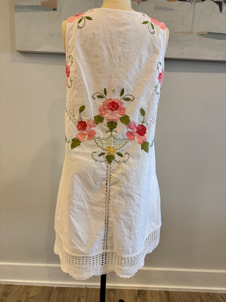 May include: White sleeveless dress with floral embroidery in pink, red, and green. The dress features a crochet lace hem and a decorative floral design on the back. The dress is displayed on a mannequin.
