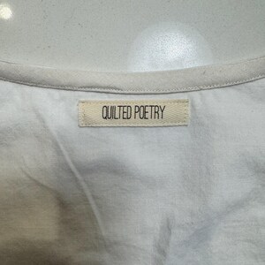 May include: Close-up of a white garment with a cream-colored trim. A rectangular label with the words "QUILTED POETRY" is sewn onto the fabric. The fabric appears to be a soft, woven material.