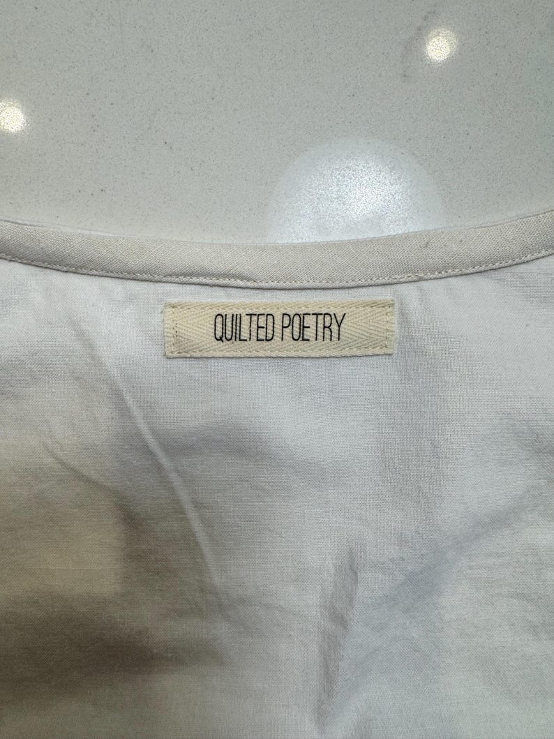 May include: Close-up of a white garment with a cream-colored trim. A rectangular label with the words "QUILTED POETRY" is sewn onto the fabric. The fabric appears to be a soft, woven material.
