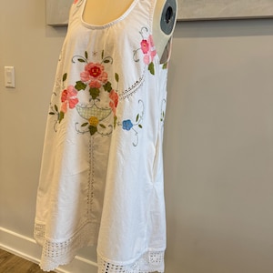 May include: White sleeveless dress with floral embroidery in pink, red, and green. The dress features a scoop neckline and a crocheted lace hem. The floral design is centered on the front, with additional embroidery along the neckline.