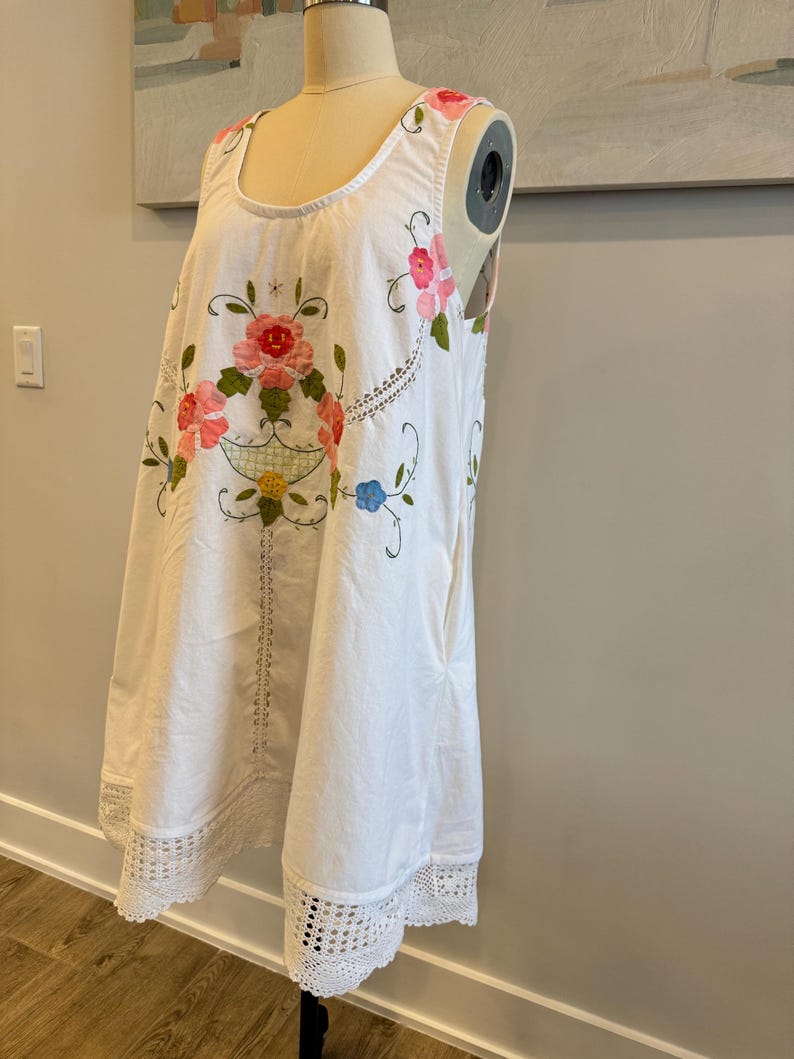 May include: White sleeveless dress with floral embroidery in pink, red, and green. The dress features a scoop neckline and a crocheted lace hem. The floral design is centered on the front, with additional embroidery along the neckline.