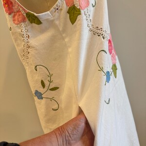 May include: White sleeveless dress with floral embroidery. The dress features pink and red flower appliqu&eacute;s with green leaves, blue flowers, and delicate vine details. The neckline and armhole are trimmed with white lace.