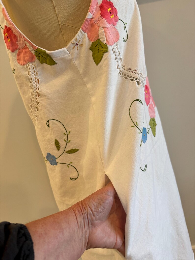 May include: White sleeveless dress with floral embroidery. The dress features pink and red flower appliqu&eacute;s with green leaves, blue flowers, and delicate vine details. The neckline and armhole are trimmed with white lace.