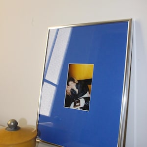 May include: A framed photograph with a silver metal frame. The photograph is set against a vibrant blue mount. The image within the frame depicts two people. A small, yellow lidded container sits in the foreground.
