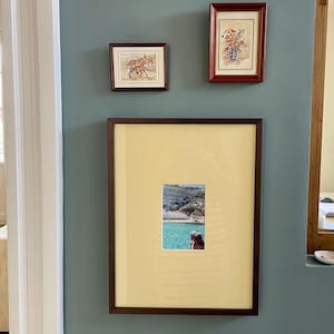 Puede incluir: Tres obras de arte enmarcadas en una pared. El marco superior izquierdo contiene una pequeña ilustración de paisaje, el marco superior derecho una pintura floral y el marco inferior grande muestra una fotografía de una persona frente a una bahía turquesa.