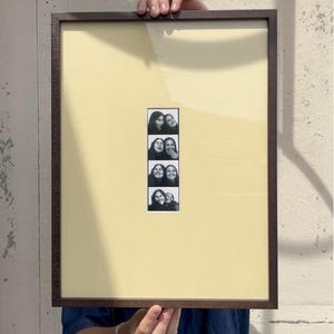Op de afbeelding: Een rechthoekige fotolijst met een donkerhouten rand en een lichtgeel passe-partout. In het midden bevindt zich een verticale zwart-wit fotostrip met vier verschillende opnames van twee lachende personen die samen poseren.