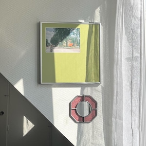May include: A square silver-framed wall art piece featuring a photograph of a street scene with trees and a vehicle, set against a solid olive green background, positioned above a small octagonal mirror with a textured pink border.