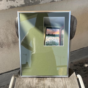 Op de afbeelding: Een ingelijste foto met een zilveren metalen lijst. De foto toont een kleinere afbeelding van een zonsondergang boven een veld met witte bloemen. De lijst heeft een groene passe-partout.