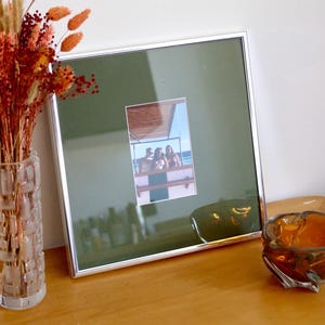 Könnte beinhalten: Ein quadratischer silberner Bilderrahmen mit einem dunkelgrünen Passepartout, das ein vertikales Foto von drei Personen auf einem Boot enthält. Links steht eine klare Glasvase mit getrockneten orangefarbenen und roten Blumen, rechts eine kleine bernsteinfarbene Glasschale.