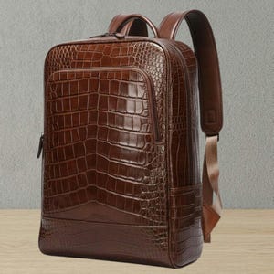 Puede incluir: Una mochila de cuero marrón con textura de piel de cocodrilo. Tiene forma rectangular, asa superior, bolsillo frontal con cremallera y correas ajustables. La mochila está hecha de cuero marrón oscuro.