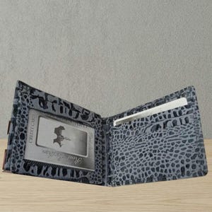 Könnte beinhalten: Offene, graue Ledergeldbörse mit reptilienartigem Muster. Die Geldbörse ist geöffnet und zeigt Kartenfächer und ein transparentes Ausweisfach. Eine Kreditkarte ist im Ausweisfach sichtbar. Die Worte "Real Leather" sind auf der ID-Karte aufgedruckt.