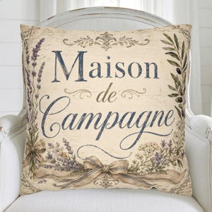 May include: A square throw pillow with a cream-colored background featuring the dark blue text Maison de Campagne. The design includes illustrations of lavender sprigs, olive branches, and a decorative ribbon bow at the bottom center.