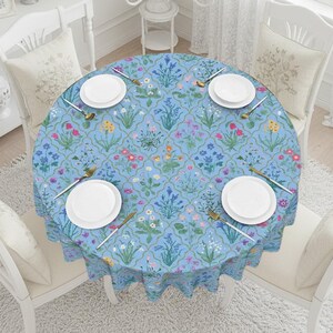 May include: A round, light blue tablecloth with a floral pattern featuring various colorful flowers and green foliage. The table is set with white plates, gold-colored utensils, and is surrounded by white chairs with floral-patterned cushions.