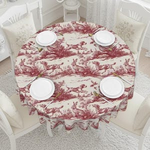 May include: A round tablecloth with a red and cream toile pattern featuring dogs and rabbits. The table is set with white plates, gold silverware, and surrounded by white chairs with matching cushions.