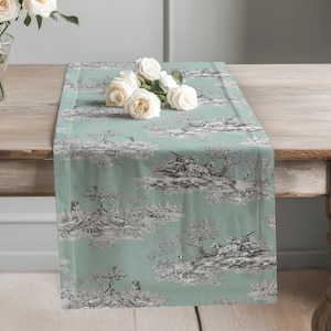 May include: A rectangular table runner in a soft sage green colour featuring a repeating black and white toile de jouy pattern of hunting dogs and birds in a woodland landscape, topped with a small cluster of white roses.