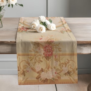 May include: A rectangular table runner featuring a beige background with a floral pattern in shades of pink, cream, and green. The design includes ornate scrollwork borders and a central bouquet of flowers, topped with a cluster of white roses.