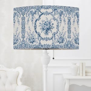 May include: A floor lamp featuring a cylindrical drum shade with a blue and white toile pattern depicting floral bouquets and ornate scrollwork. The lamp has a white pedestal base and a black pull chain switch hanging from the bottom edge.