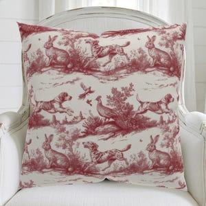 May include: A decorative pillow with a red toile pattern featuring rabbits, dogs, and birds in a pastoral scene. The pillow is square and rests on a white chair. The design is a classic, detailed illustration.