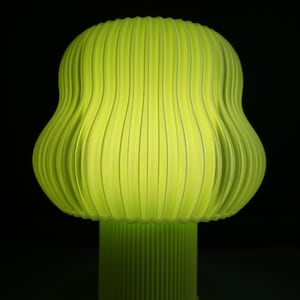 Könnte beinhalten: Eine limettengrüne Lampe mit geripptem, abgerundetem Schirm und zylindrischem Sockel. Die Lampe hat eine strukturierte Oberfläche und strahlt sanft vor dunklem Hintergrund. Das Design ist modern und skulptural, geeignet für die Inneneinrichtung.