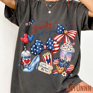 May include: Dark gray t-shirt with a colorful graphic featuring patriotic and Disney-themed elements. The design includes a bow, high heel, popcorn, and a drink, with the text "Literally just a girl".