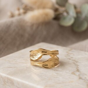 May include: A gold-toned ring featuring an open, double-band design with a textured, wavy surface. The ring consists of two parallel, curved bands that are connected at the back, creating a fluid, organic shape with a polished, metallic finish.