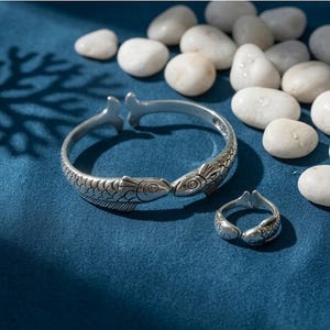 Könnte beinhalten: Ein silberfarbenes, offenes Armband und ein passender offener Ring, beide mit einem detaillierten Fischdesign mit strukturierten Schuppen und Flossen. Die Schmuckstücke sind mit einem oxidierten Finish versehen, um die Fischköpfe zu betonen.