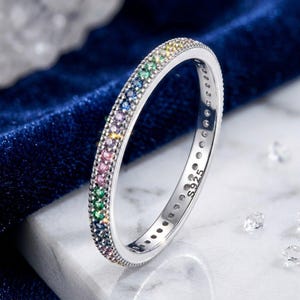 May include: A thin sterling silver ring featuring a channel of small, multi-colored gemstones in shades of green, blue, purple, and pink. The inner band is clearly stamped with the text S925, indicating the metal composition of the jewelry piece.