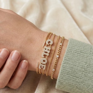 May include: A set of four gold-toned bracelets worn on a wrist. One bracelet features the name Chloe spelled out in clear pavé stones, while another is a curb chain with three evenly spaced clear stones. Two additional thin gold bangles are included.