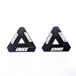 May include: Two black Crocs shoe charms in a triangular shape. Each charm has the word "Crocs" in white lettering on each side of the triangle. The charms are set against a white background.