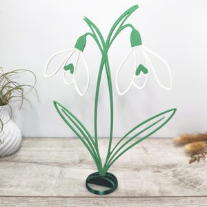 Peut inclure: Sculpture décorative de fleurs de perce-neige en métal vert et blanc. La sculpture représente deux fleurs de perce-neige avec des pétales blancs et des accents verts, des tiges et des feuilles vertes. La base est un ovale vert foncé.